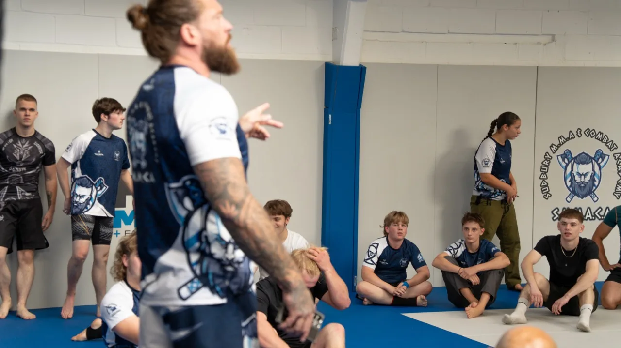 A coach addressing his class of students seated on the mats in the gym